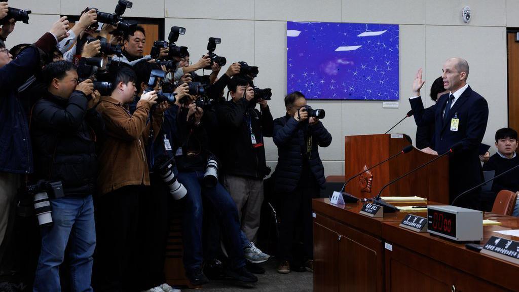 Harold Rogers from Coupang is seen holding his hand up to take an oath during a hearing into a data breach while a bank of photographers take images.