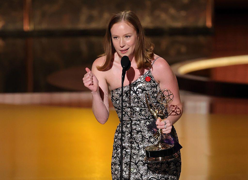Hannah Einbinder wears a sparkling gown with a red pin affixed to it as she speaks into a microphone onstage at the Emmy Awards. She holds her Emmy trophy for supporting actress in a comedy in her left hand.