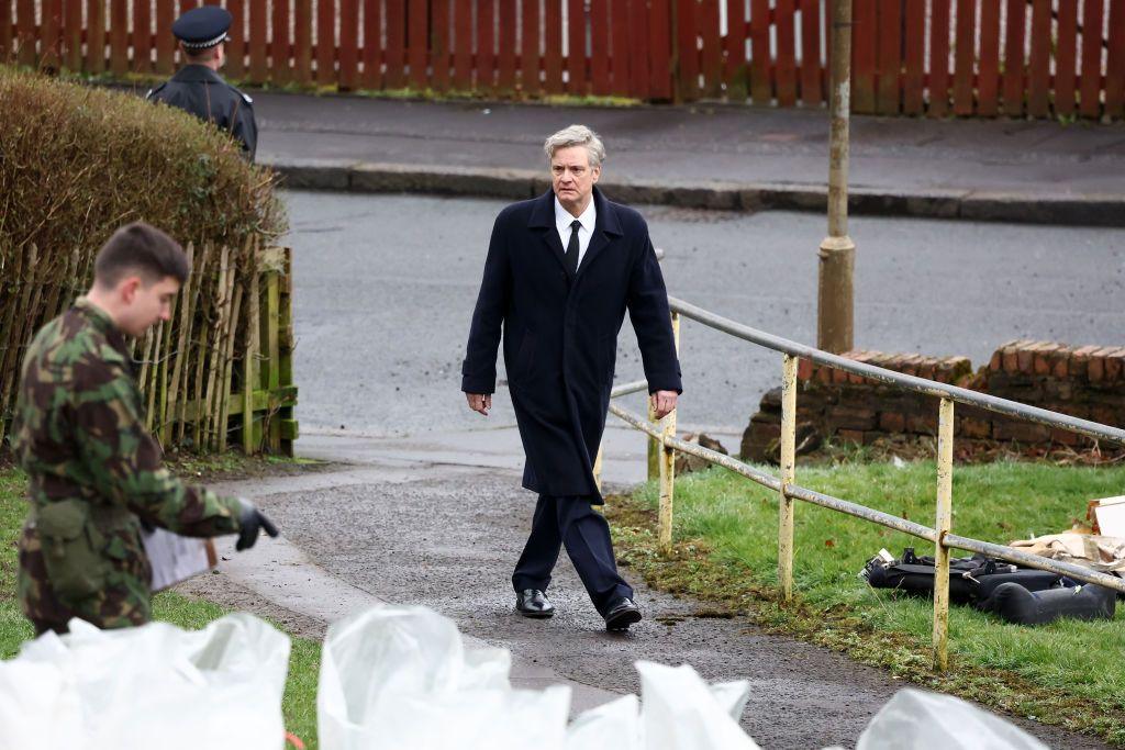 Colin Firth on set in character as Dr Jim Swire - he is walking up a pathway looking concerned an is wearing a long dark coat, a white shirt and a black tie. There is an army officer in the foreground and a police officer in the background.