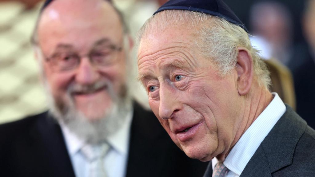 King Charles III, wearing a grey suit with a pink pocket square, wears a blue kippah while meeting the congregation at the Heaton Park Synagogue
