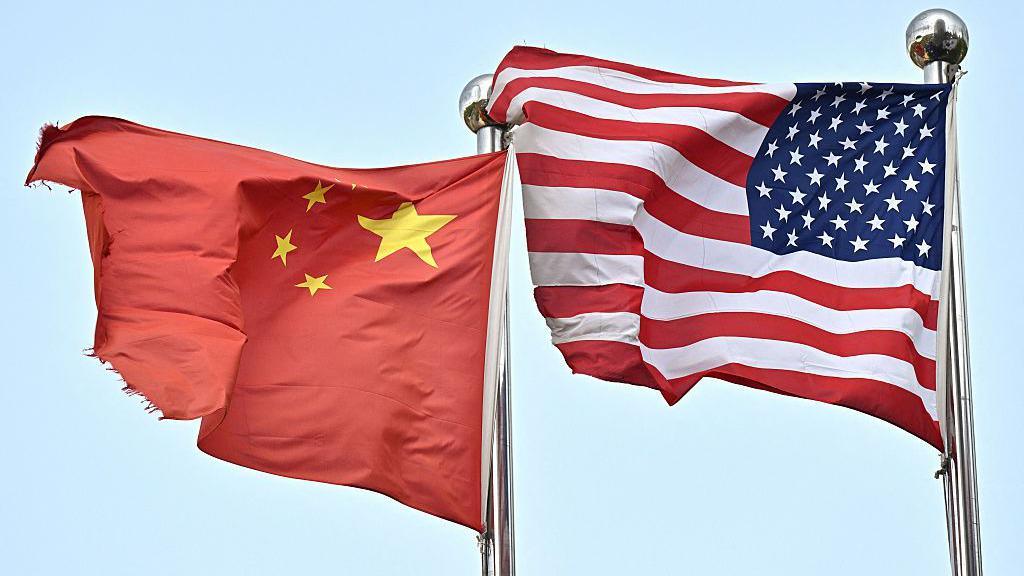 Chinese flag blowing in the wind on the left and US flag blowing in the wind on the right. Both are mounted on flag poles