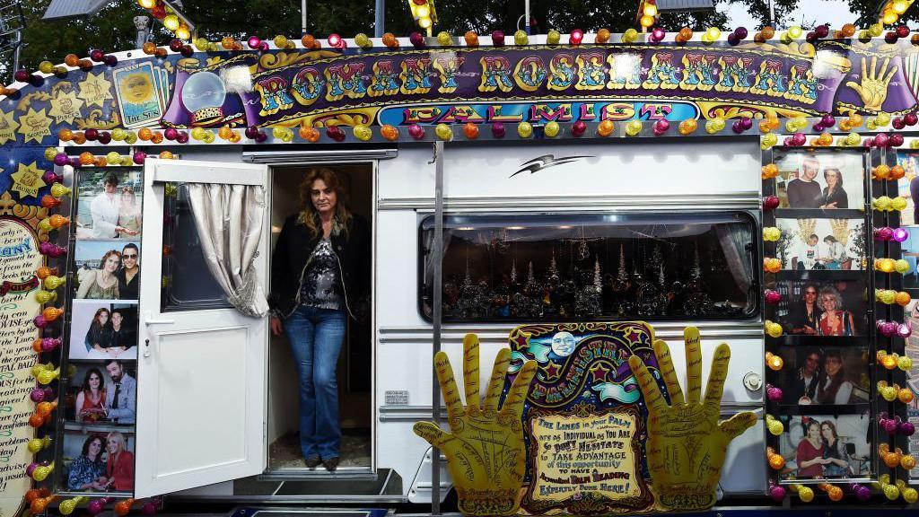 A woman stands in the door of a caravan. She is wearing blue jeans and a black jacket and the caravan is covered in lights and has the words Romany Rosehannah along the top.