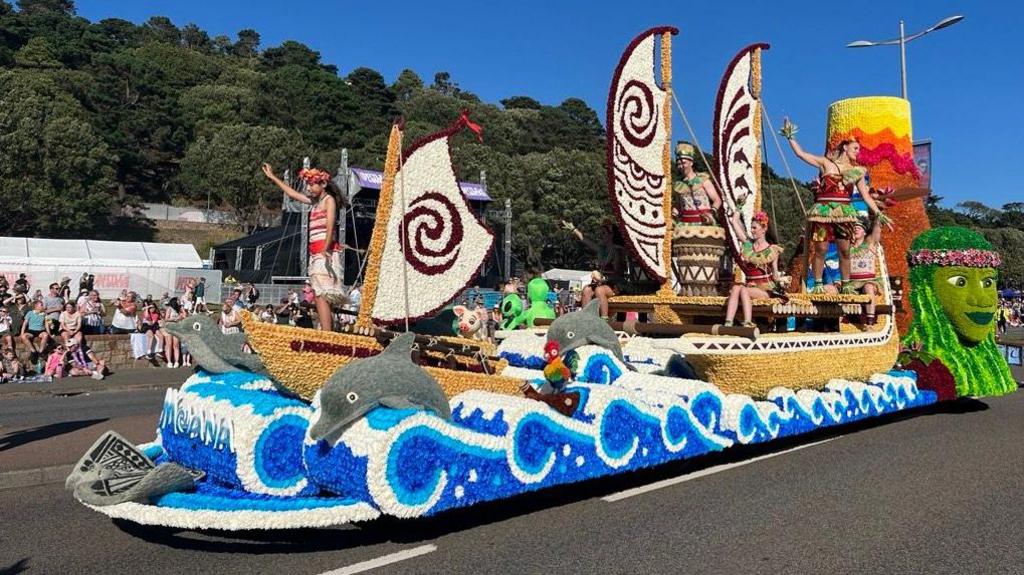 A brightly coloured float with waves and dolphins made from flowers. people on the float are dressed in traditional Hawaiian clothing