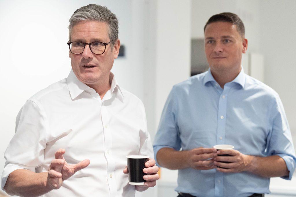 Prime Minister Sir Keir Starmer, mid-speech and wearing a white shirt, standing in front of Health Secretary Wes Streeting, who's wearing a blue shirt. Both are holding black cups.