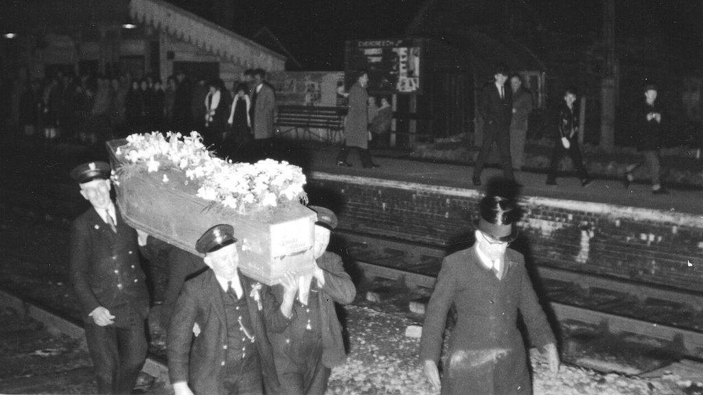 Train drivers carrying the coffin on the tracks, with a crowd of people watching on the platform.