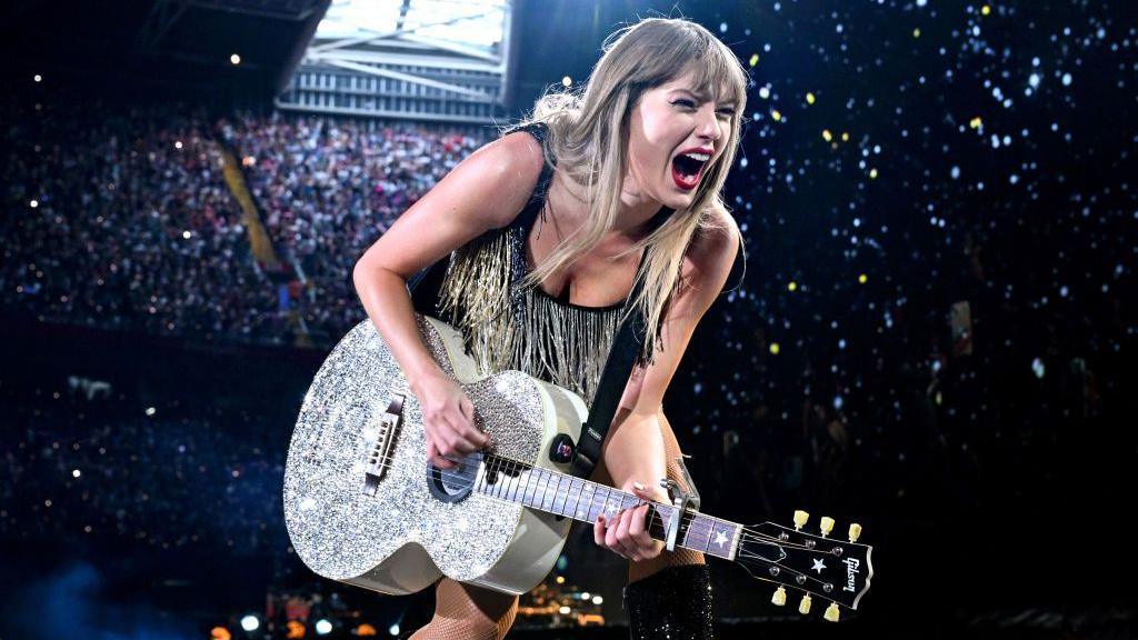 Taylor Swift performing on stage. She is wearing a black and gold sparkly dress and is holding her silver guitar. She is looking out to the audience with dramatic facial expressions and her mouth open. Behind Swift are people sitting in the stands. The roof is closed.