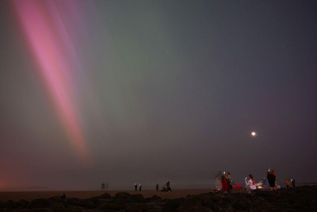 People stand in open fields at night filming the sky with phones as pink rays of the northern lights flood down from the top left corner into the middle of the frame.