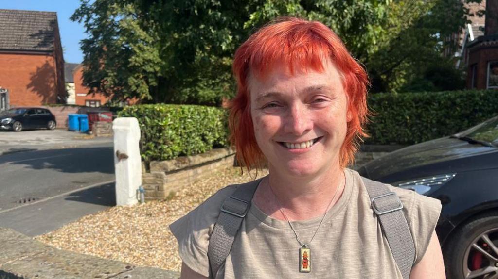 Tracey Cartledge, who has short, cropped red hair and is wearing a light brown t-shirt and backpack, smiles while standing on the pavement in front of a driveway.