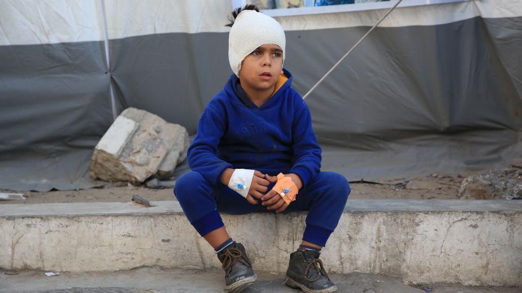 A Palestinian boy sits on the sidewalk with his head wrapped in bandages and an IV in his hands at Al-Ahli Baptist Hospital