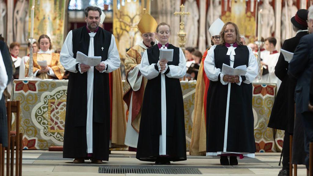 The Venerable Barry Hill, who was ordained Bishop of Whitby, the Venerable Flora Winfield, who was ordained Bishop of Selby, and the Venerable Tricia Hillas standing next to each other holding hymn sheets in York Minster.