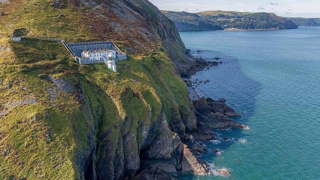 Foreland Point Lighthouse in Exmoor National Park. It is a large white building with a stone wall behind it, separating it from the moorland. In front of it is the white lighthouse with a wraparound balcony. It is situated very close to the cliff edge, where below, waves break around the jagged rocks. The water is a bright blue and in the background there is another headland stretching out into the sea.