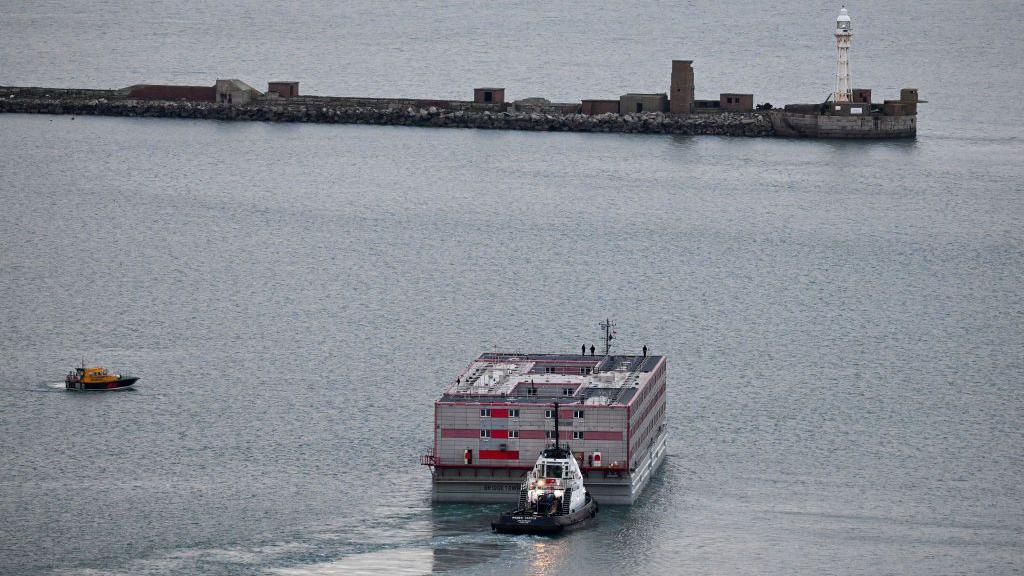 The Bibby Stockholm moves through the water away from Portland. The barge is red and white and there is a white tugboat behind it and a yellow and black smaller boat the the left. 