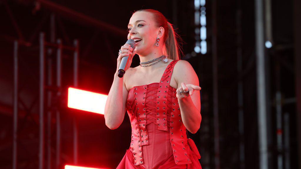 A photo of a woman singer on a stage. She is in an all red corset with her hair up in a ponytail. She is also being illuminated by a red stage light.