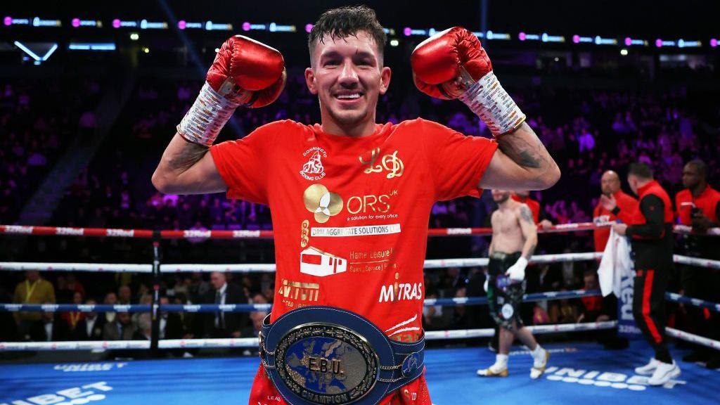 Liam Davies is posing in a blue boxing ring. He is smiling and is wearing red boxing gloves with his arms raised in the air. He is wearing a red t-shirt and a large blue boxing trophy belt.