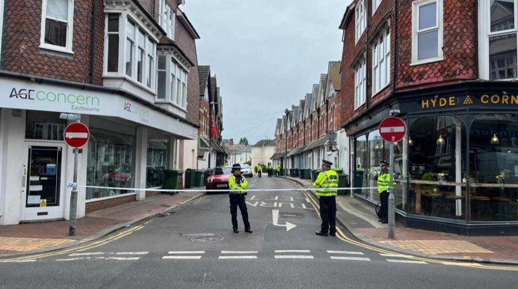 Police stand by a cordoned-off road. It is daylight.