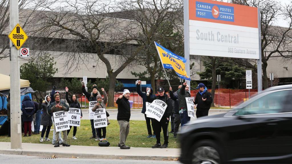 People stand on a sidewalk and hold signs as they cheer at passing cars. There is a sig above them that reads Canada Post South Central