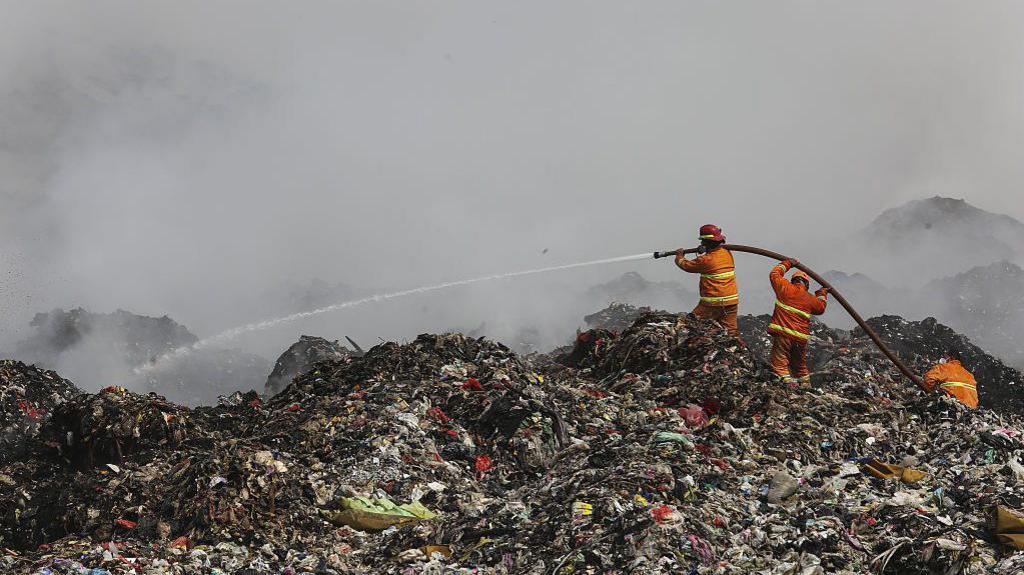 Firefighters in orange uniforms standing on a gigantic pile of rubbish, holding a hose and spraying water, as smoke billows into the air