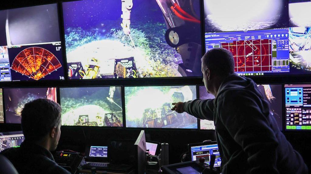 Two men looking at a number of screens showing scenes from under the sea