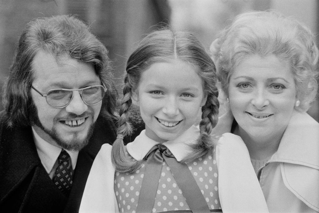 Black and white photo of a young Lena Zavaroni standing in between her parents, Victor on the left and Hilda on the right. Her hair is in pigtails and she is wearing a spotted dress. Victor, who has glasses on, has a 70s-style haircut and a jacket and tie. Hilda is in a light jacket and pearl earrings and they are all looking at the camera.