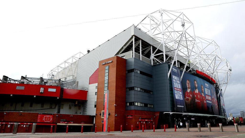 The outside of Manchester United's Old Trafford. It is silver and red, with large white metal protruding from the top of it. There is a large black screen above its entrance with photos of players on it.