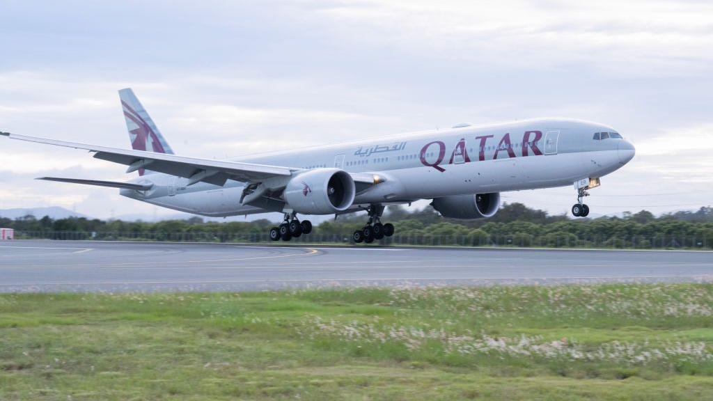 A wide shot of a plane taking off on a runway. "Qatar" is printed on the body of the plane.