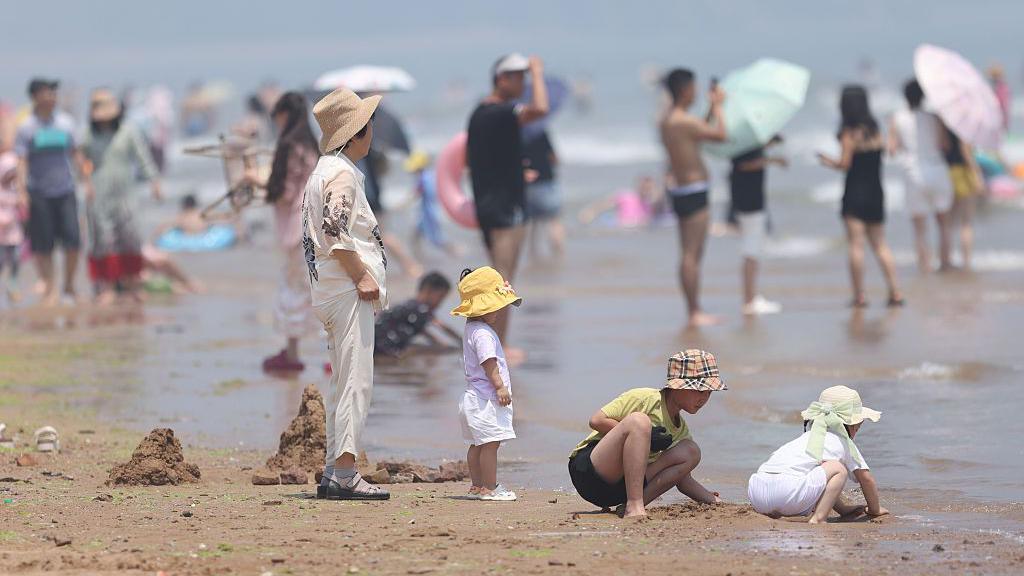 A middle-aged woman wearing a brown hat standing on a beach. In front of her are three children playing with sand. In the background are many people on the crowded beach.