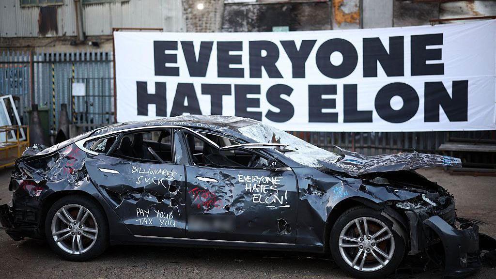 A banner reading "everyone hates Elon" behind a smashed up Tesla