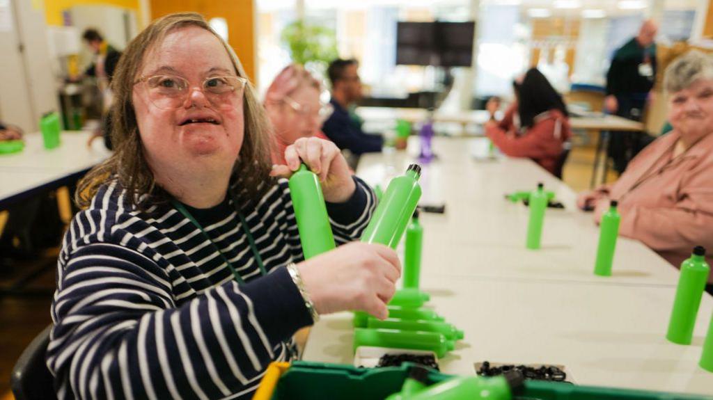 Adults at MK Snap, holding green bottles. The woman close to the front is wearing a black and white striped top, with a green lanyard round her neck. She is wearing glasses and is smiling. There are other people slightly blurred in the background. The woman is sitting by a long white table. 