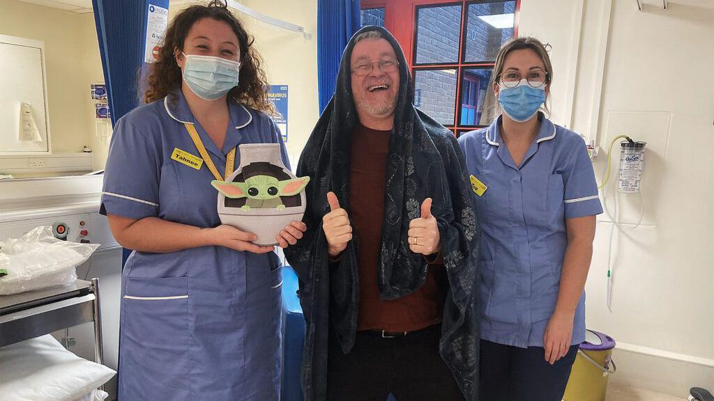 Mr Hale in hospital with two nurses beside him, while he was recovering from bowel cancer. He is smiling and giving two thumbs up for the camera. He is dressed as a Jedi and one of the nurses is holding a baby Yoda hot water bottle. 