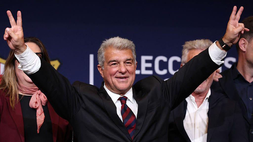 Joan Laporta raises his hands in celebration after winning the Barcelona presidency election. He has grey hair and is wearing a dark suit, white shirt and a tie in Barcelona's club colours - red and blue.