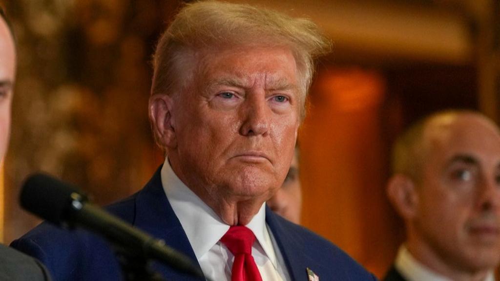 A close up shot of Trump's face, not smiling, as he stands behind a microphone at a news conference at Trump Tower