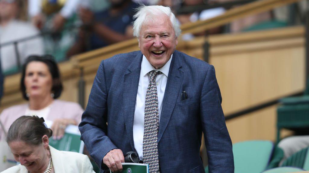 Sir David Attenborough at Wimbledon. He is smiling as he takes his seat, and is wearing a dark blue-grey jacket and and black and white patterned tie