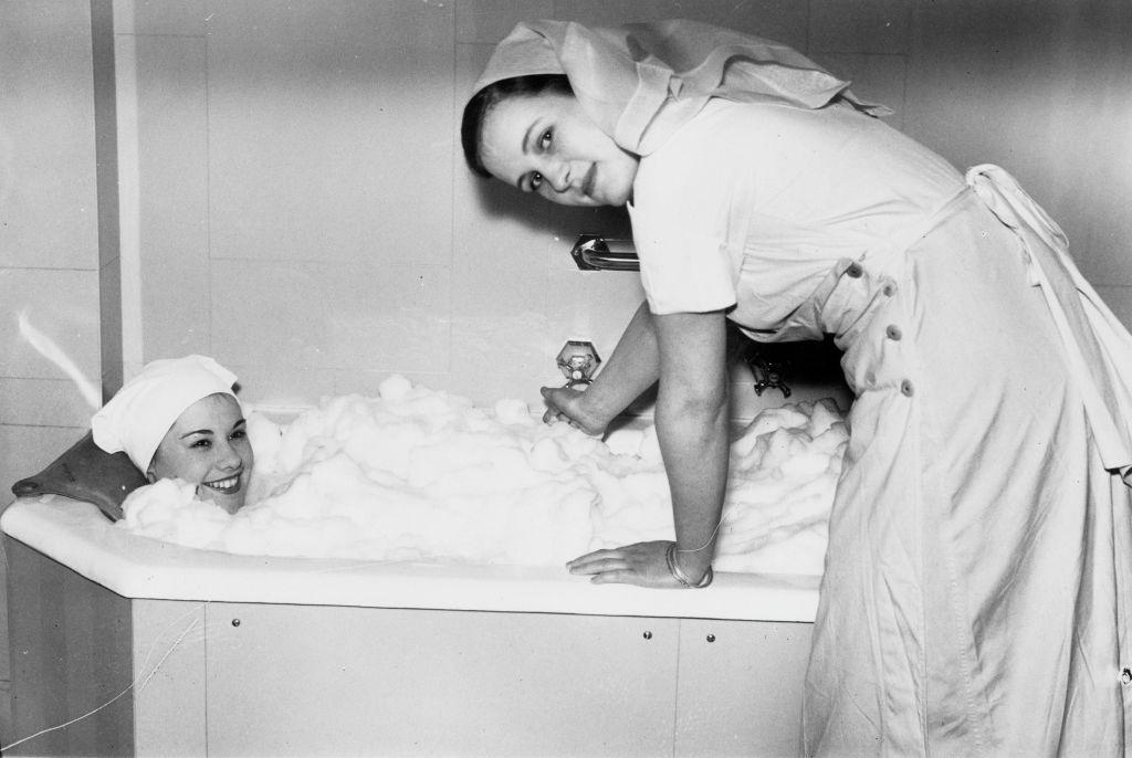 Black and white image of a woman in a bubble bath with a wrap on her head and a beautician dressed as a nurse swilling the bubbles around