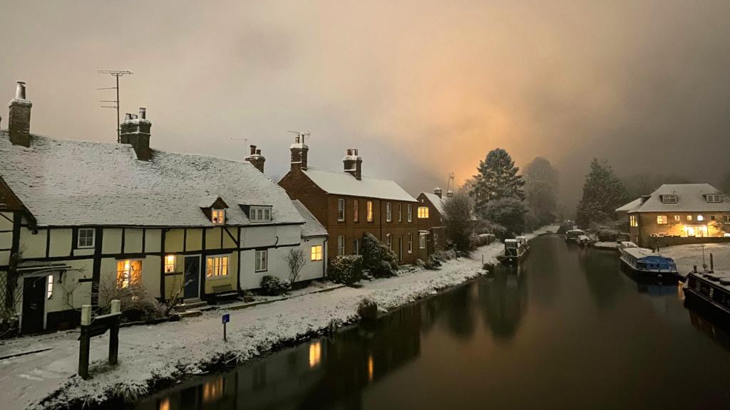 Snow lines a footpath beside a canal next to a Tudor-style home in West Berkshire. Canal boats can be seen and there is a glow of lights on the horizon, but it is night time. It is overcast.