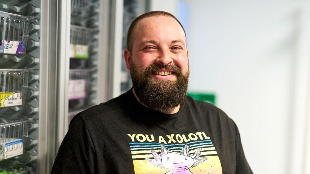 Joe - a man with short brown hair and a beard - grins at the camera. To his left you can see some of the pharmacy lab. He's wearing a black graphic t-shirt.