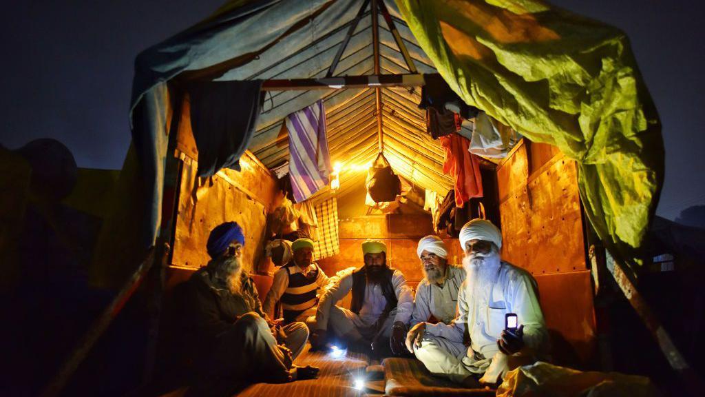 A group of turbaned men seen sitting in a makeshift shelter at the back of a tractor trolley, with a yellow light glowing in the background.