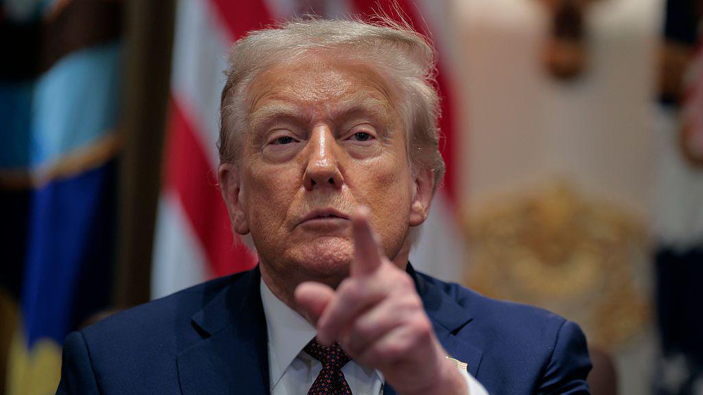 Donald Trump pointing his finger at reporters, wearing a suit and red tie, during an Oval Office meeting at the White House in late August.