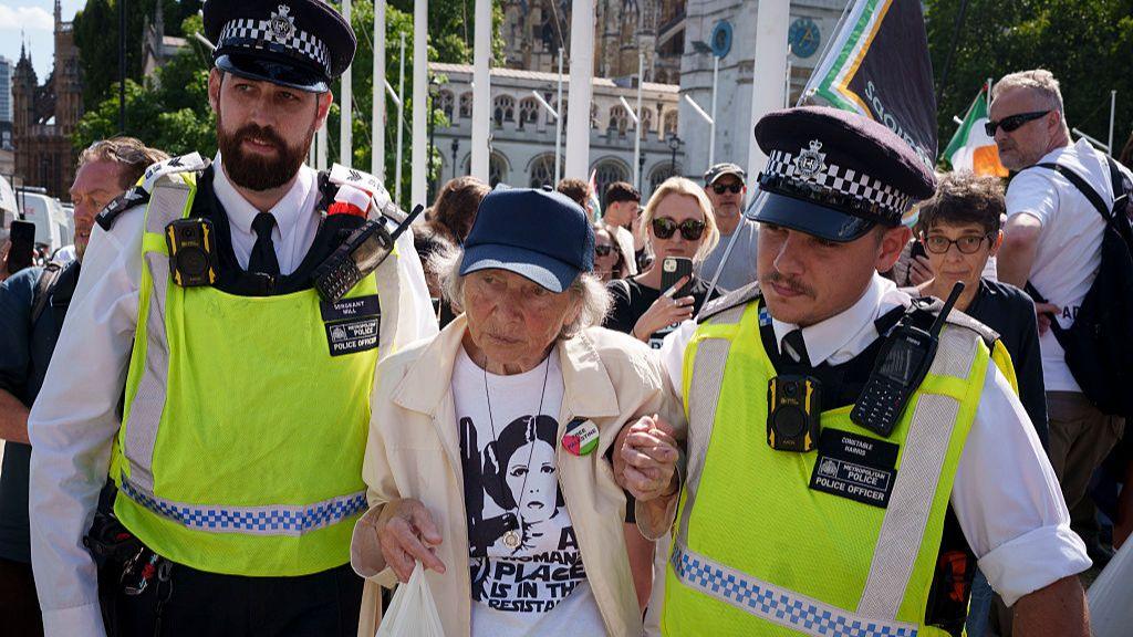 An elderly woman is led away from protesters by two police officers who flank her, with one appearing to be clutching her hand.