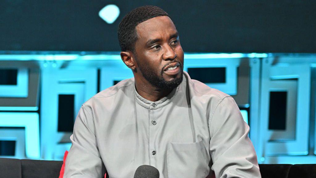 Sean "Diddy" Combs is photographed during an interview. He is sitting in a tan button-down shirt and holding a microphone. 