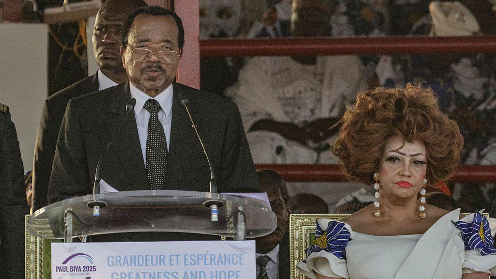President Paul Biya stands at a lectern and speaks into a pair of microphones. He is flanked by his wife, Chantal Biya.