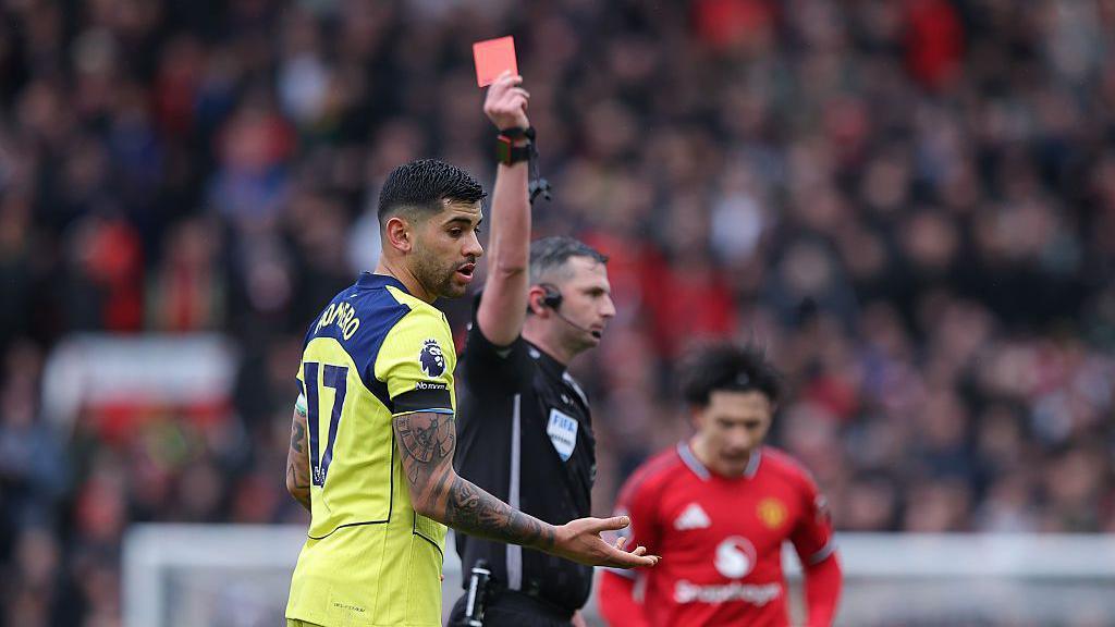 Cristian Romero reacts as he is shown a red card by referee Michael Oliver. Romero has short black hair is wearing Spurs' yellow away kit. Oliver has short grey hair and is weaing all-black. His right arm is raised as he issues the red card.