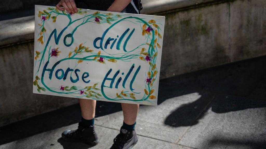 A sign which reads No drill Horse Hill being held by an unseen person, it is decorated with pictures of flowers