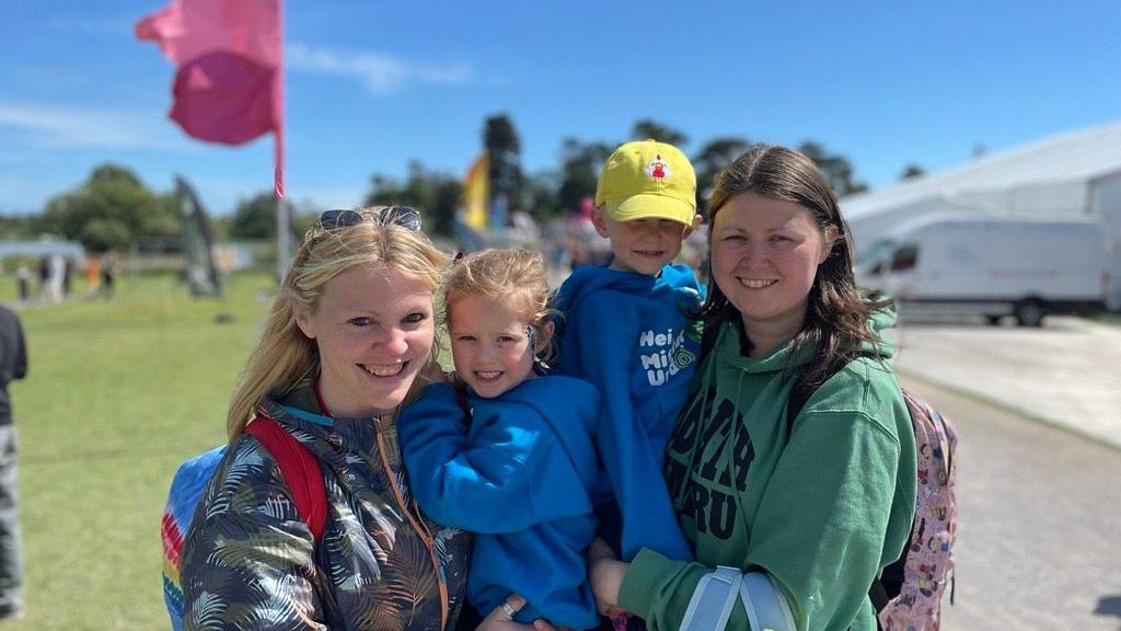 Two women hold two young children, aged five, wearing blue hoodies. One child wears a bright yellow cap. Flags can be seen in the background, and the sun shines on their faces.  One woman on the left has a blue, orange and white coat, and has blonde hair, and is smiling at the camera. The other has a green hoody and dark brown hair. 