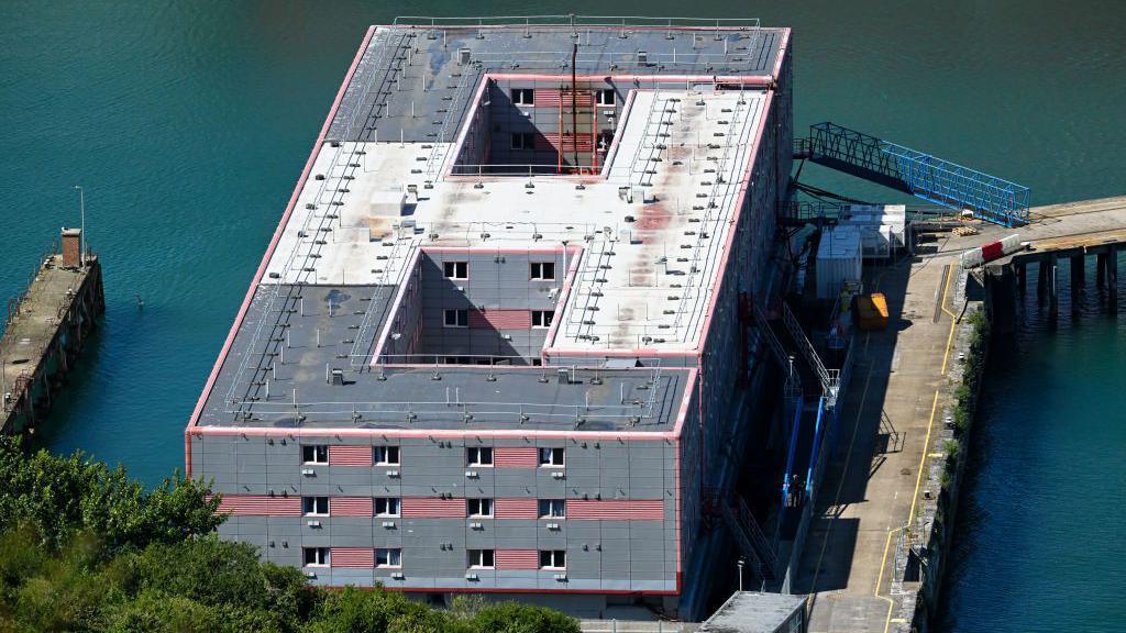 Bibby Stockholm, a grey, rectangular barge moored against a dock. It shows 12 windows.