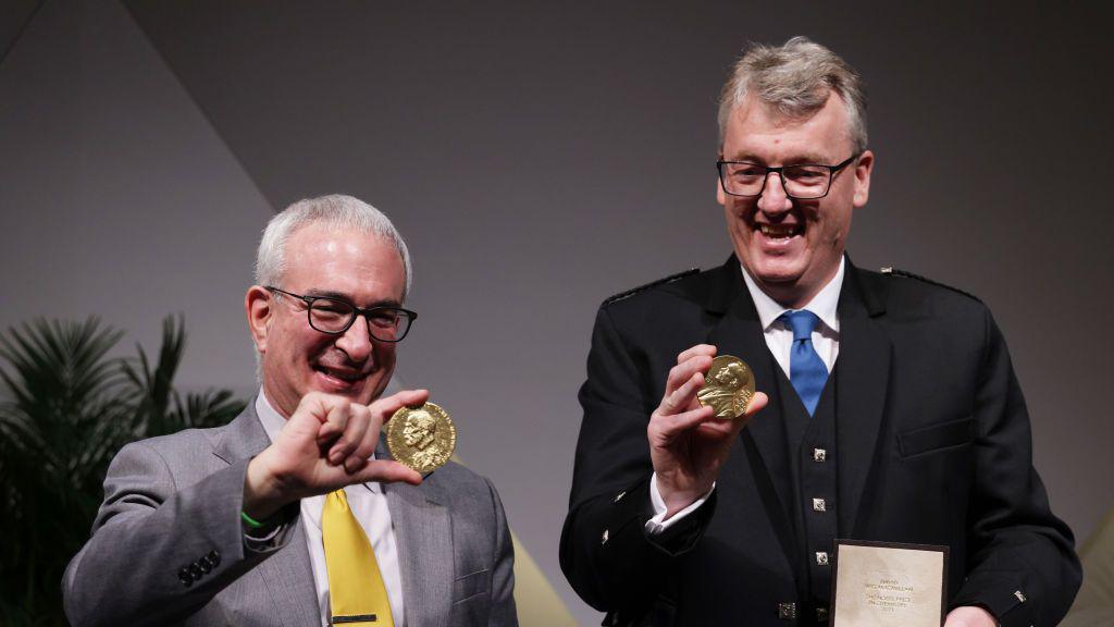 Benjamin and David both hold their Nobel Prize medals. David smiles and is on the right of the image wearing a blue tie. Benjamin smiles on the left of the image wearing a yellow tie with a grey suit. 