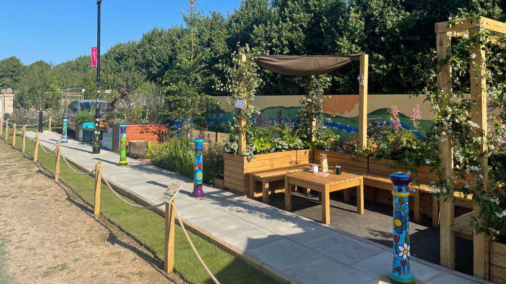 Wooden bench seating in a U shape in an area the size of a standard parking space. There is also a small wooden table and green plants behind the back of the seats. Along the path are other gardens filled with green plants.
