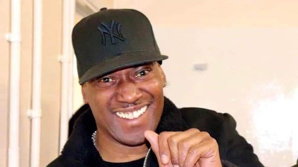 Robert Powell wearing a black cap and a black jacket and smiling at the camera. He is wearing a chain around his neck.