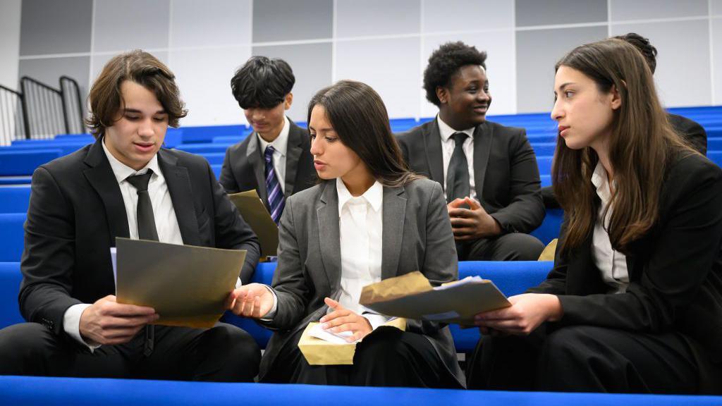 Six students sit on blue seats looking at a set of documents.