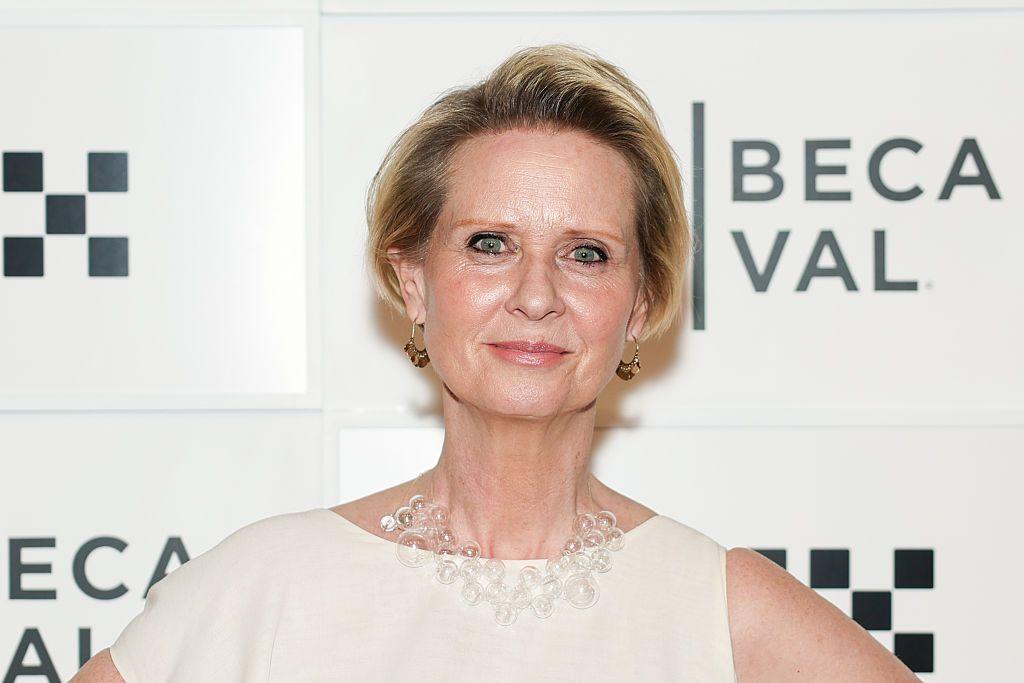 Cynthia Nixon stands in front of a sign wearing a white gown