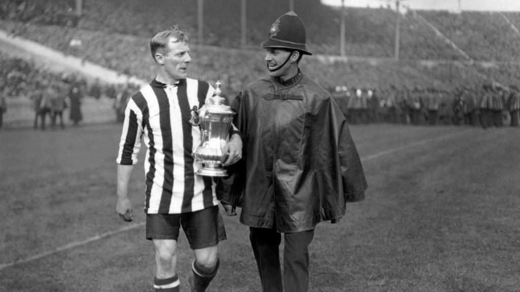 Newcastle United captain Frank Hudspeth is escorted from the pitch with the FA Cup trophy after Newcastle United's 2-0 victory over Aston Villa in the FA Cup final at Wembley Stadium.